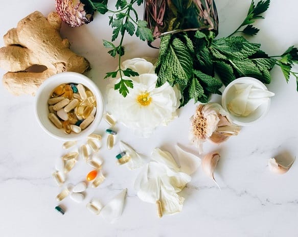 Supplement capsules in a dish surrounded by fresh herbs, flowers, and ginger