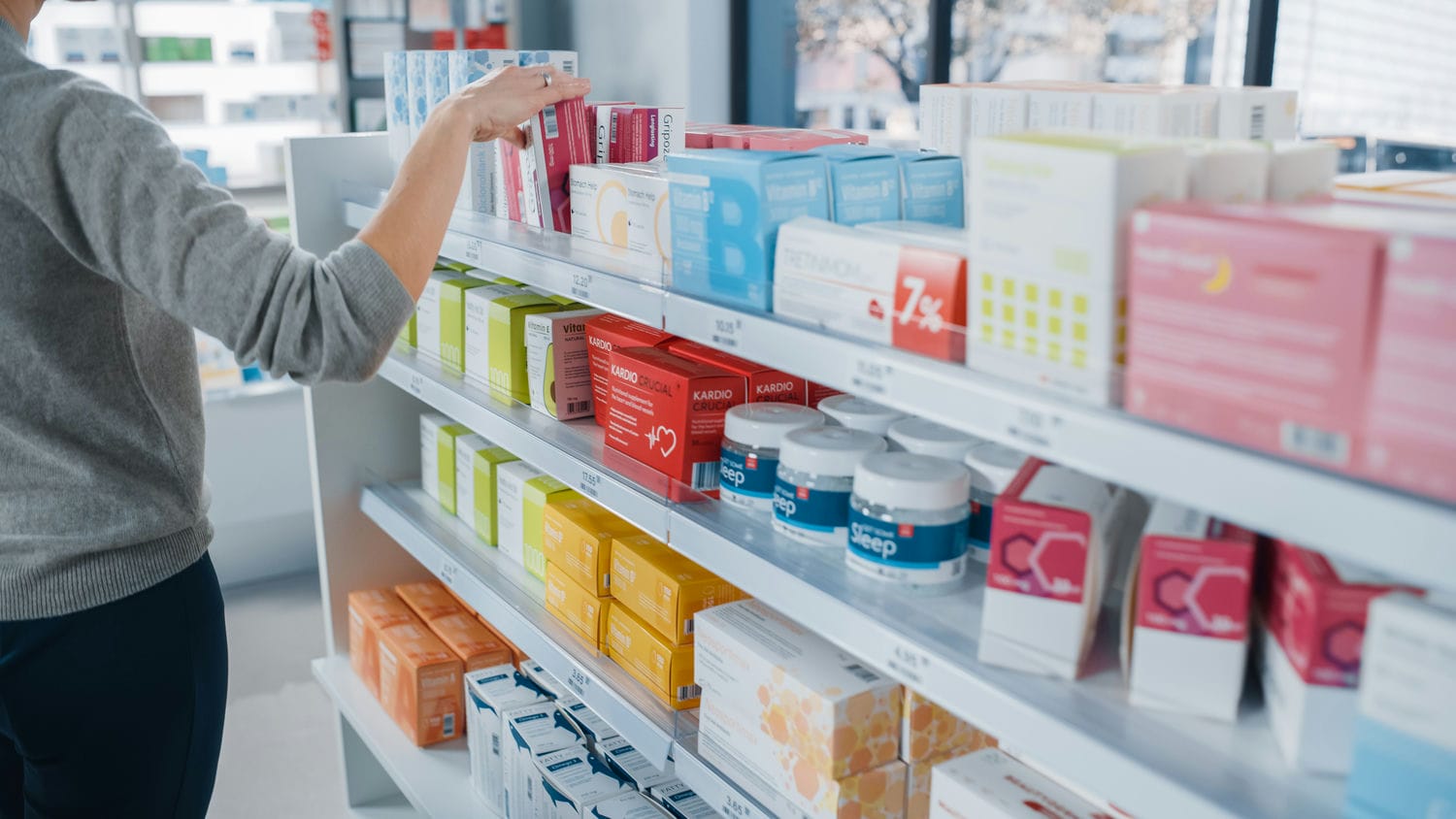 Customer reaching for an over-the-counter medicine box on a stocked pharmacy shelf